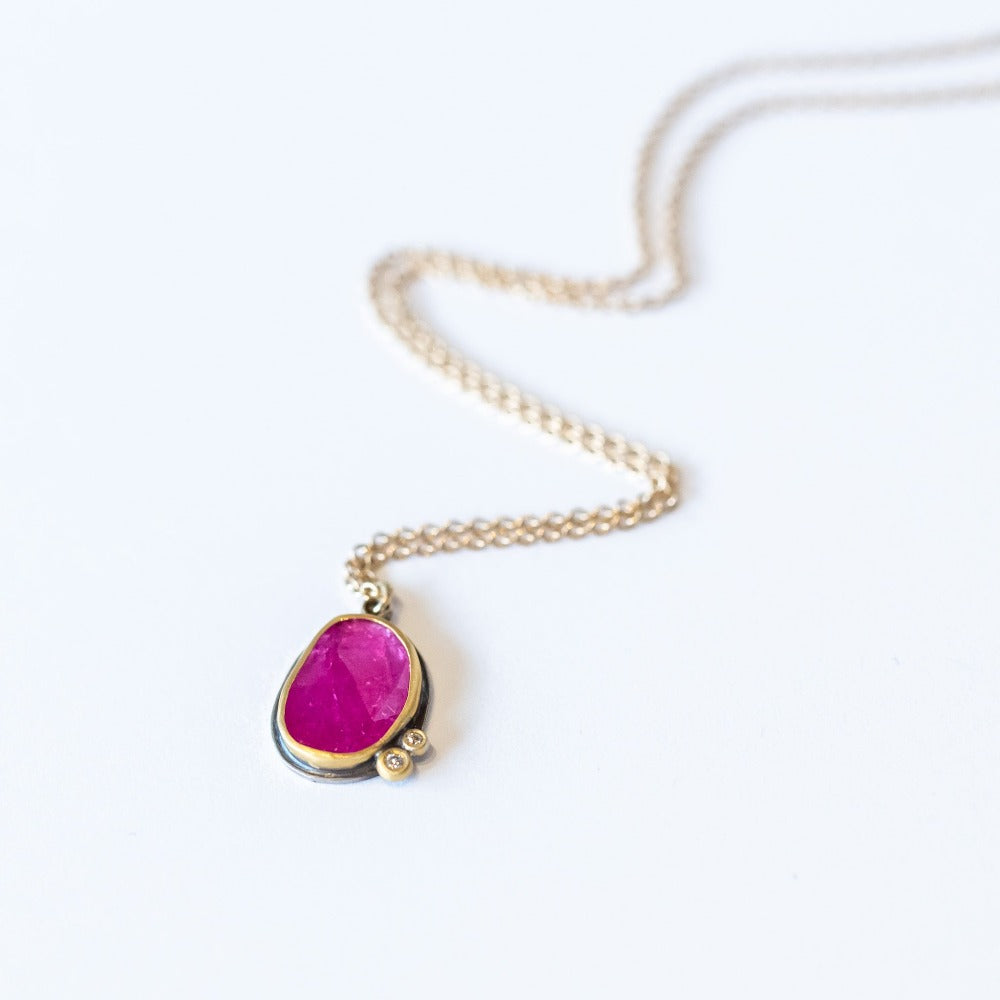 A bright pinkish-red, rose cut ruby is set with two tiny white diamond accents in 22k yellow gold bezels backed in oxidized sterling silver on a yellow gold chain necklace.
