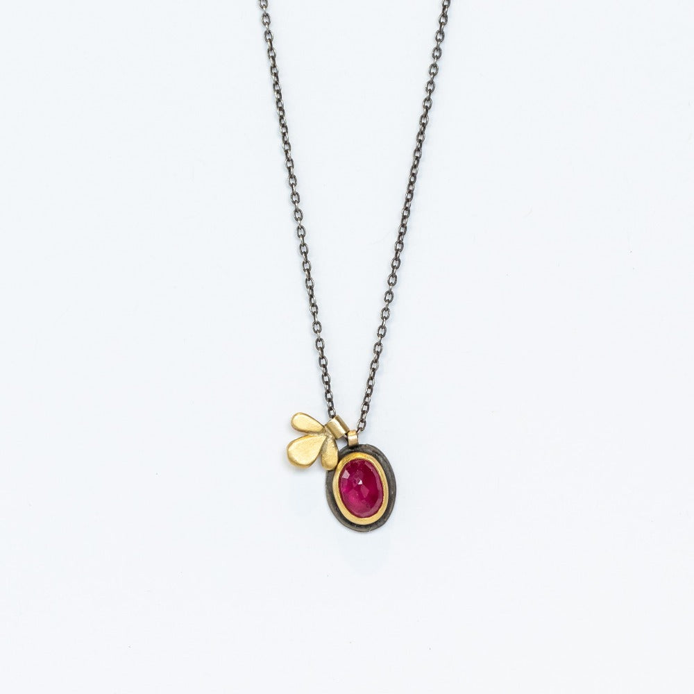 An oval ruby is bezel set in yellow gold and blackened silver, accompanied by a tiny gold leaf charm and strung from an oxidized silver necklace.