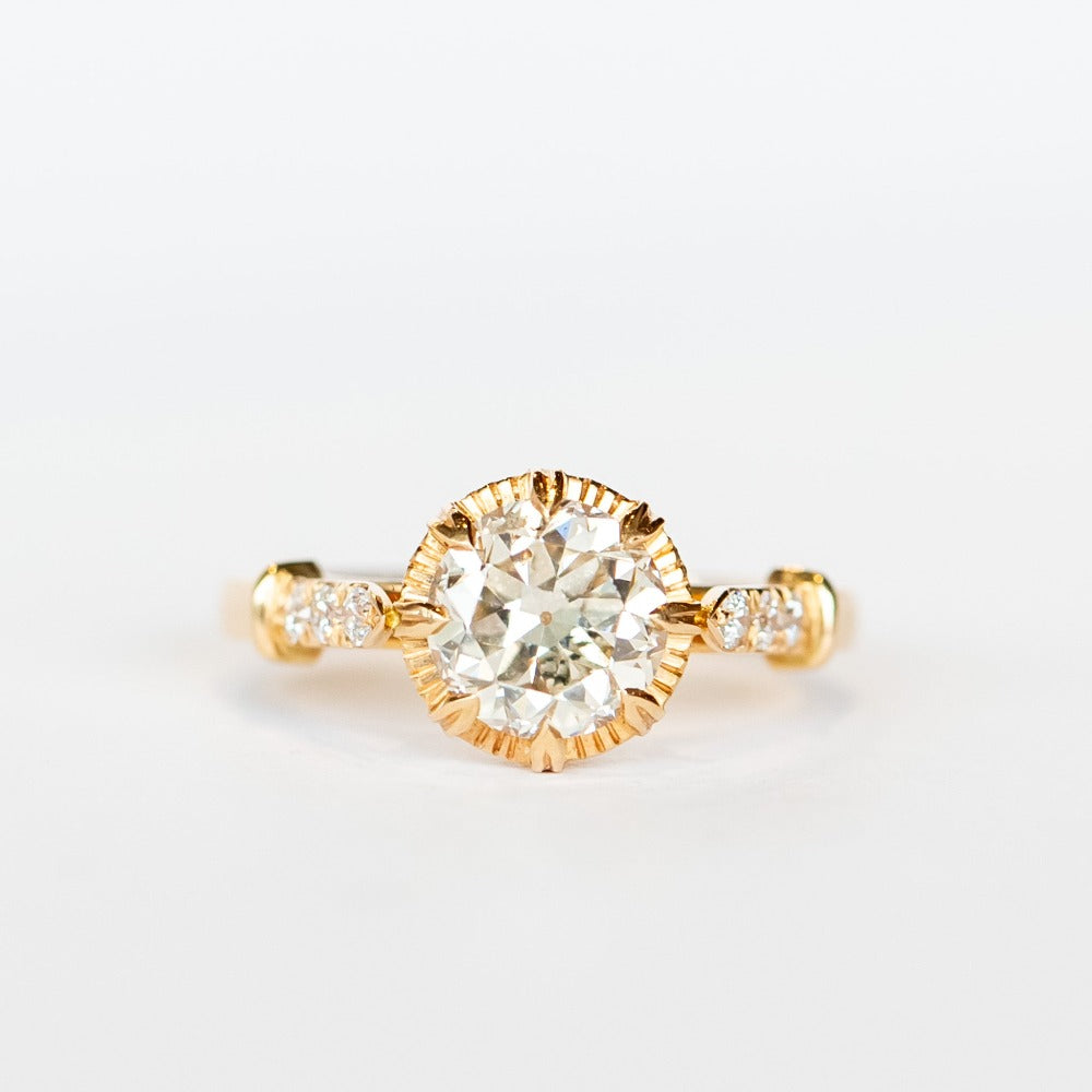 A diamond engagement ring with a round white center stone and 8 prong setting with vintage detailing. Each cathedral shoulder of the gold ring has three tiny accent diamonds.