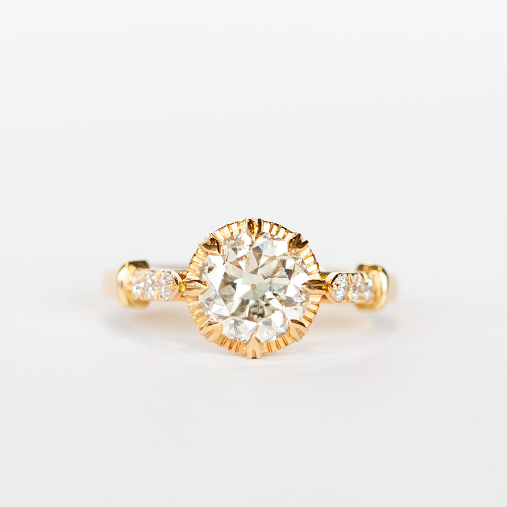 A diamond engagement ring with a round white center stone and 8 prong setting with vintage detailing. Each cathedral shoulder of the gold ring has three tiny accent diamonds.