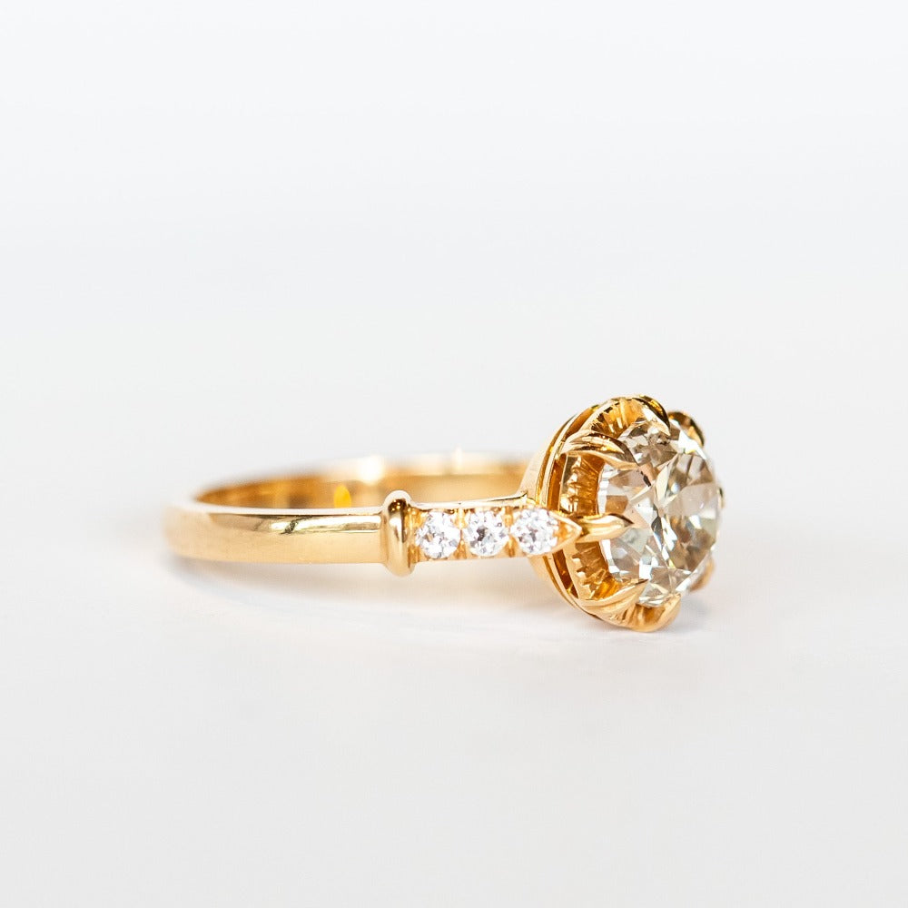 A diamond engagement ring with a round white center stone and 8 prong setting with vintage detailing. Each cathedral shoulder of the gold ring has three tiny accent diamonds.