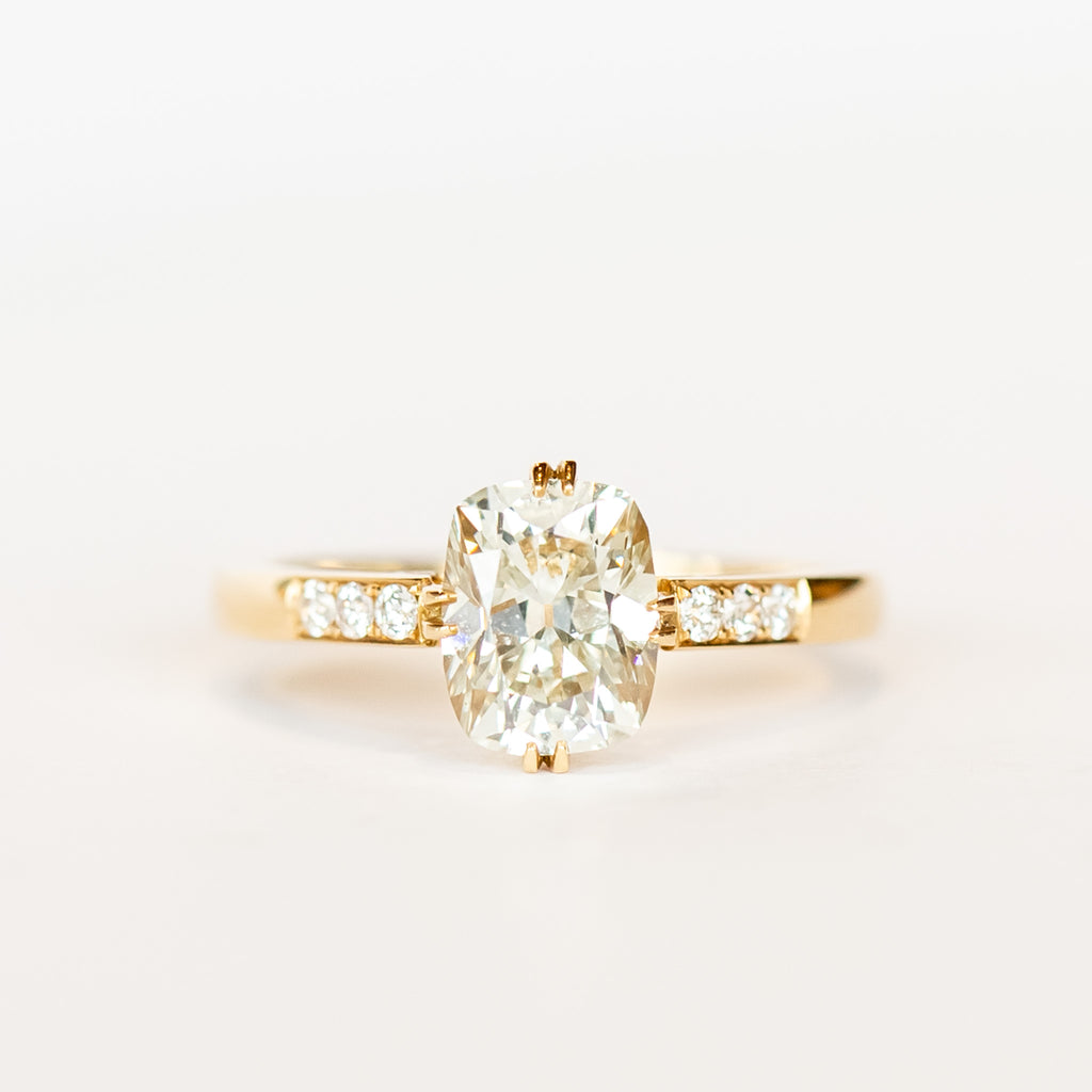 A yellow gold engagement ring set with a cushion cut diamond center stone in compass point double claw prongs, flanked by three small diamonds on each shoulder.