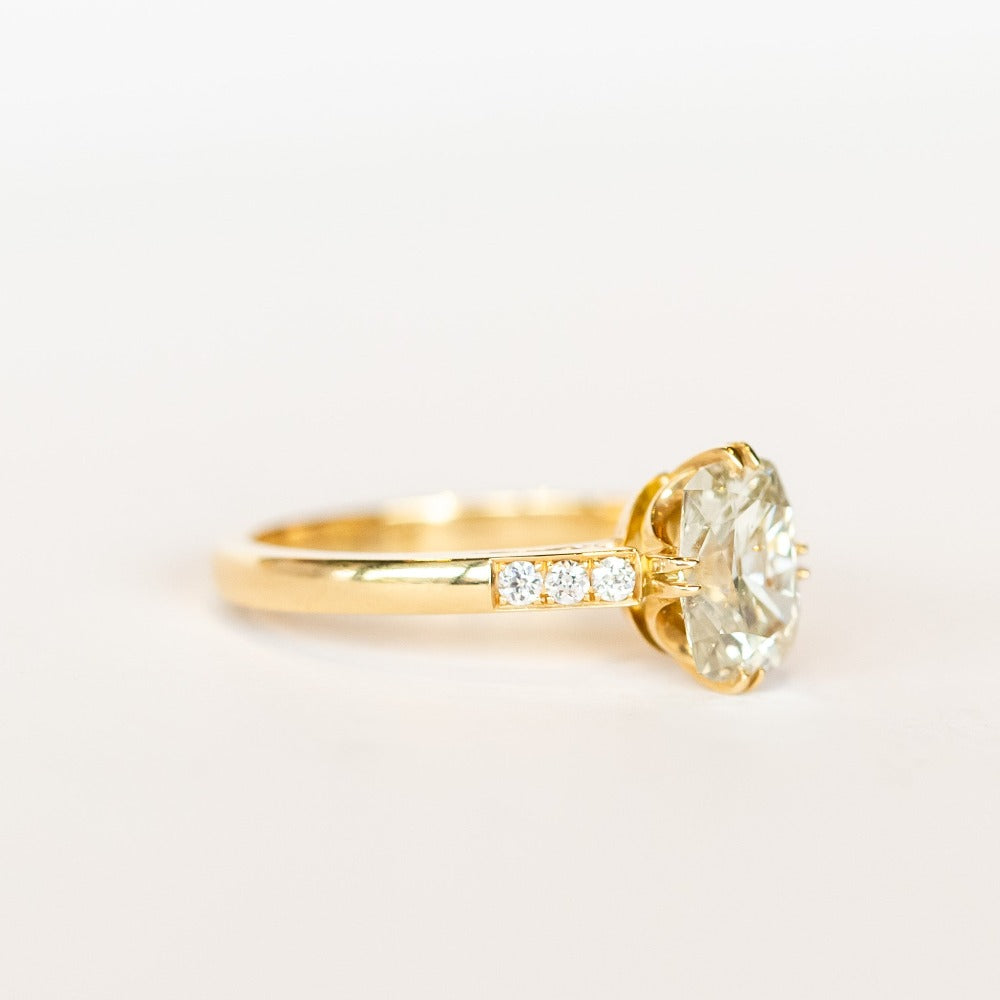 A yellow gold engagement ring set with a cushion cut diamond center stone in compass point double claw prongs, flanked by three small diamonds on each shoulder.