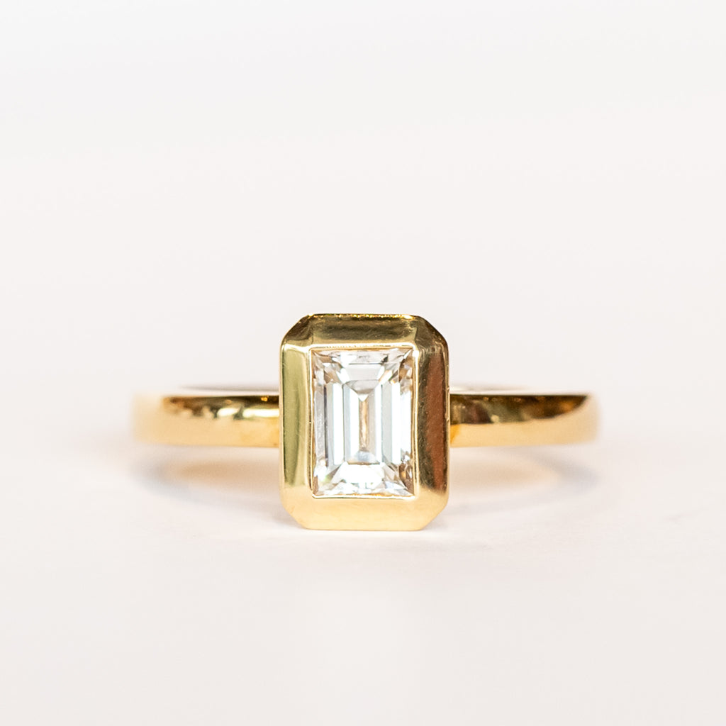 A yellow gold solitaire engagement ring featuring a bezel set rectangular step cut diamond.