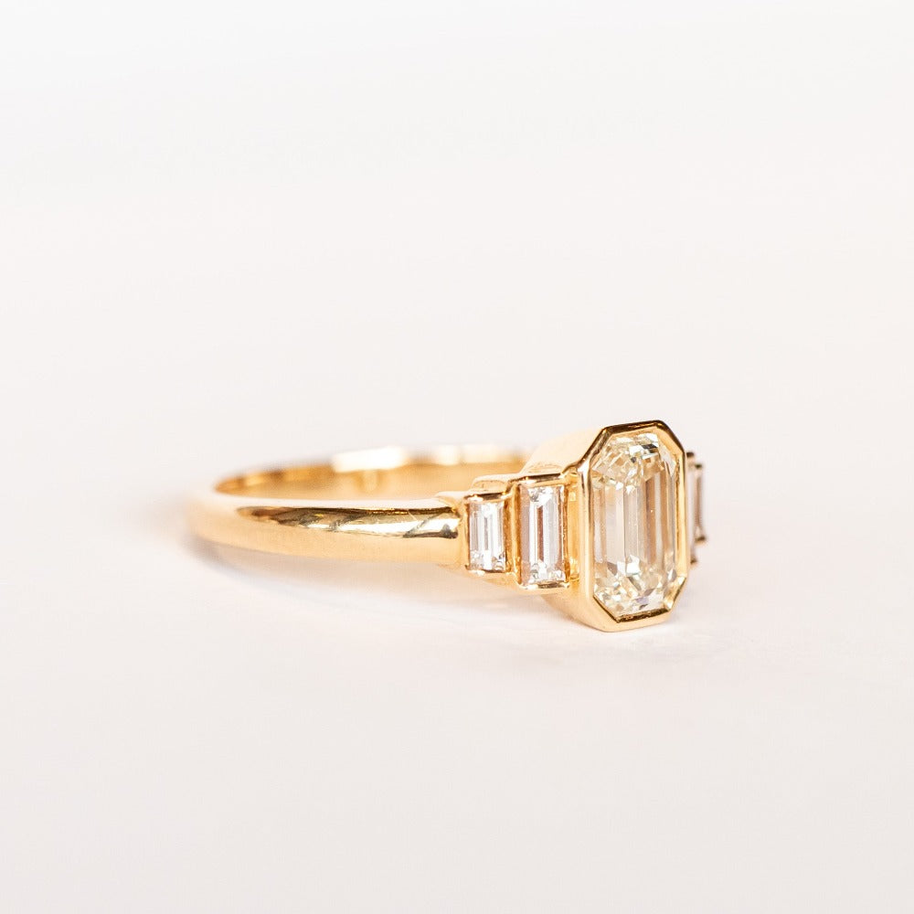 A rose gold engagement ring featuring a bezel set emerald cut diamond center stone flanked by two vertically set baguette diamonds in graduating sizes on either side.