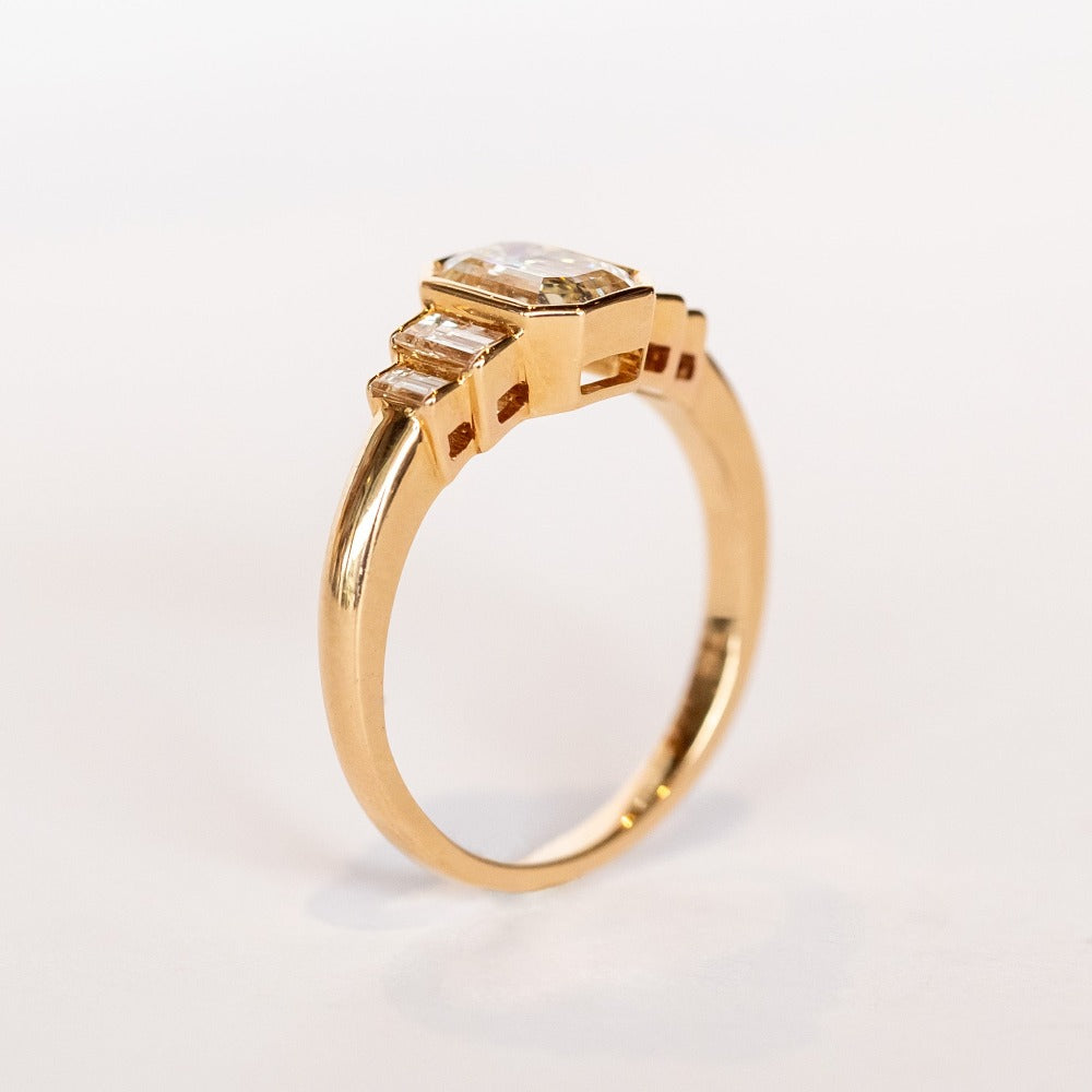 A rose gold engagement ring featuring a bezel set emerald cut diamond center stone flanked by two vertically set baguette diamonds in graduating sizes on either side.