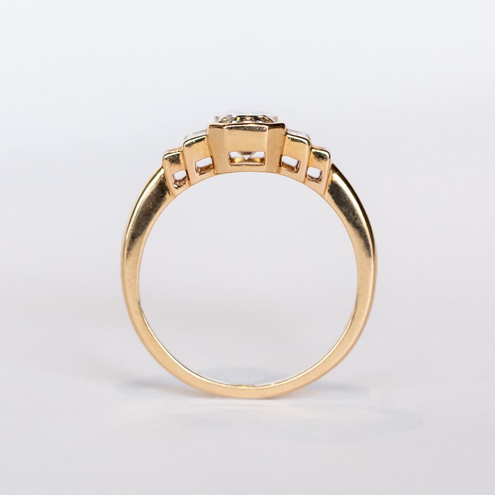 A rose gold engagement ring featuring a bezel set emerald cut diamond center stone flanked by two vertically set baguette diamonds in graduating sizes on either side.