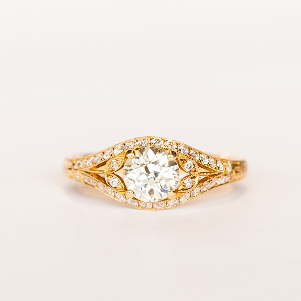 A yellow gold engagement ring that features an old European cut center diamond, floral filigree accents, and rows of pave diamonds.