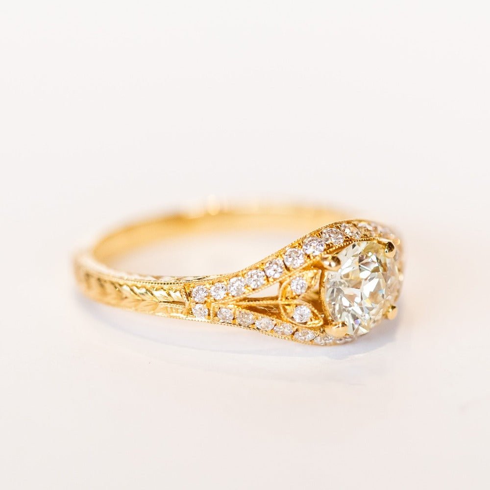 A yellow gold engagement ring that features an old European cut center diamond, floral filigree accents, and rows of pave diamonds.