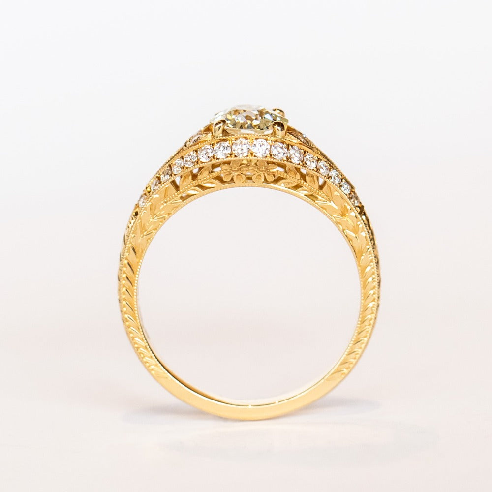 A yellow gold engagement ring that features an old European cut center diamond, floral filigree accents, and rows of pave diamonds.