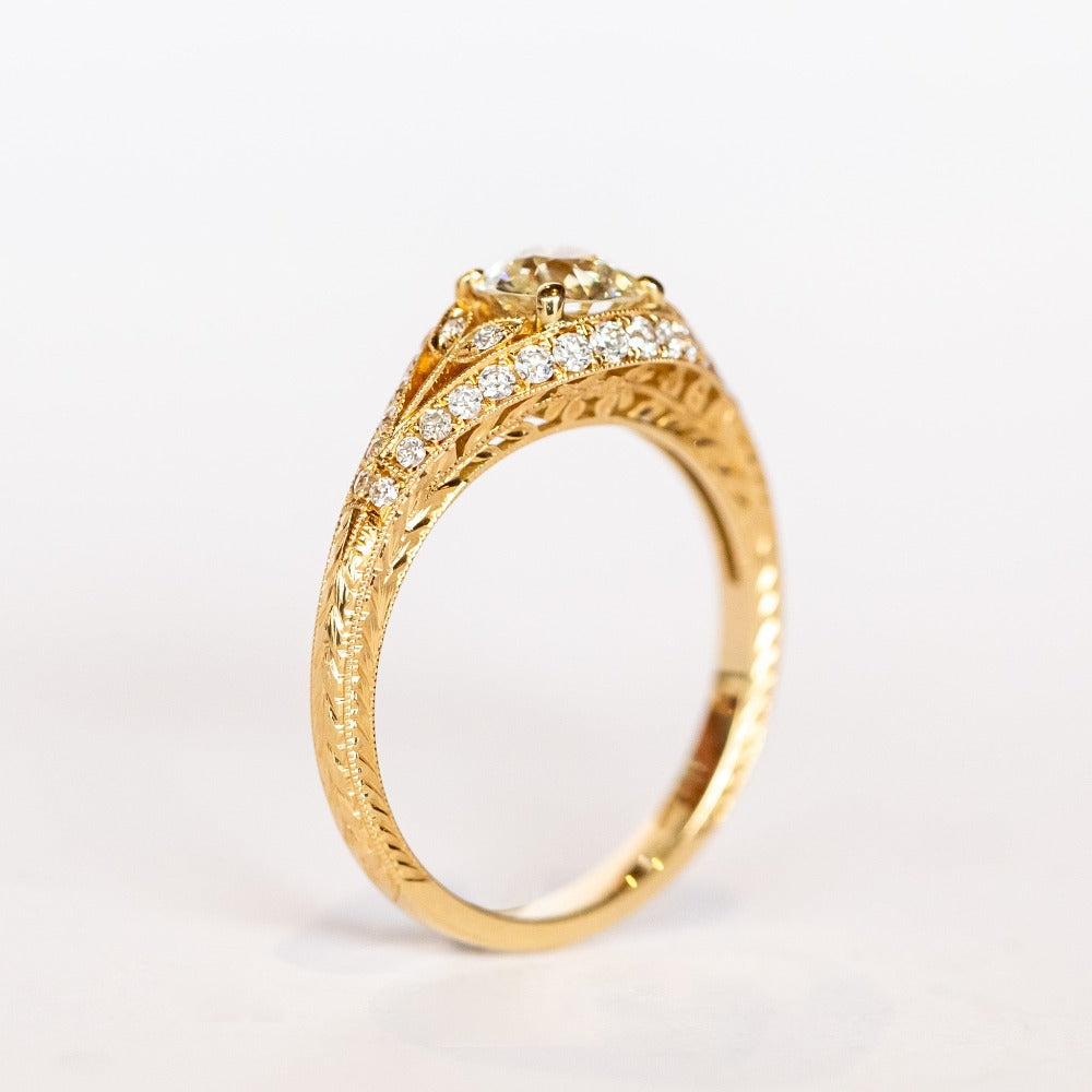 A yellow gold engagement ring that features an old European cut center diamond, floral filigree accents, and rows of pave diamonds.