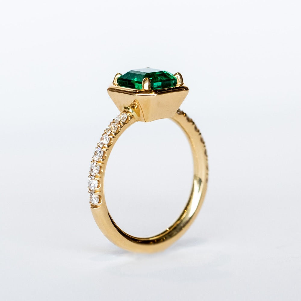 An engagement ring with an Asscher cut center emerald gemstone in yellow gold with accent diamonds on each shoulder of the ring.