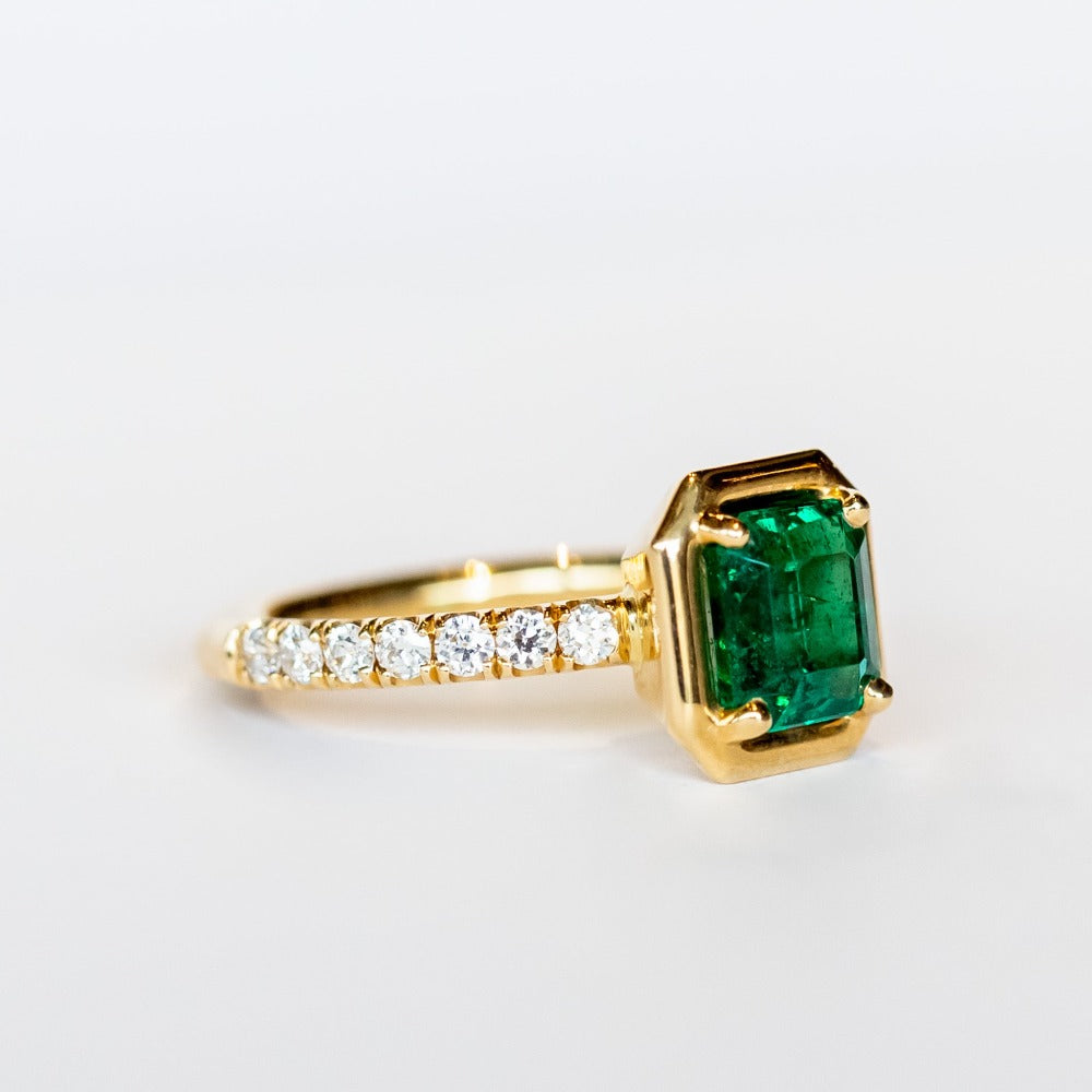 An engagement ring with an Asscher cut center emerald gemstone in yellow gold with accent diamonds on each shoulder of the ring.
