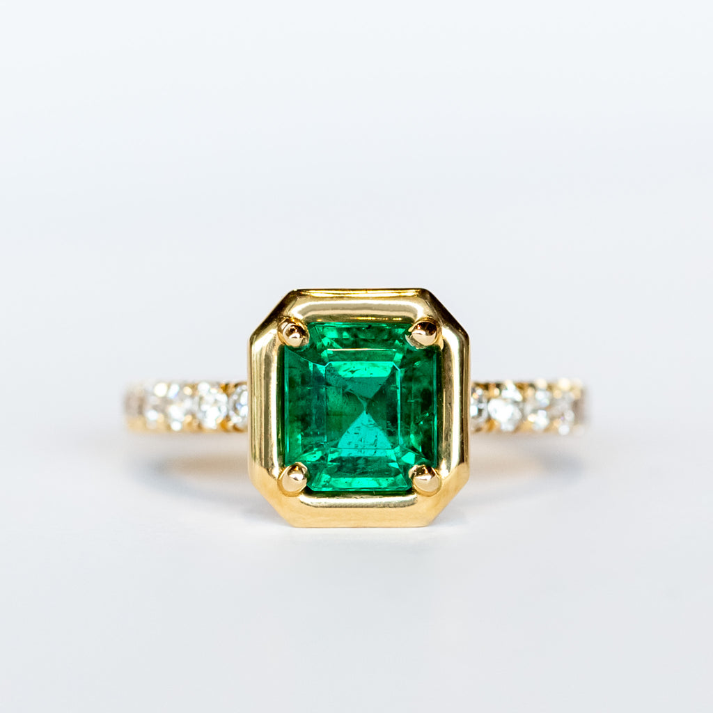 An engagement ring with an Asscher cut center emerald gemstone in yellow gold with accent diamonds on each shoulder of the ring.