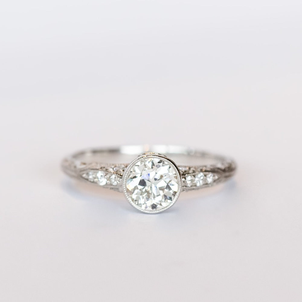 A platinum diamond engraved engagement ring with a bezel set old mine cut diamond center stone and a few diamond accents on each shoulder.