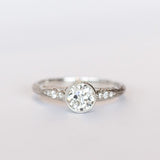 A platinum diamond engraved engagement ring with a bezel set old mine cut diamond center stone and a few diamond accents on each shoulder.