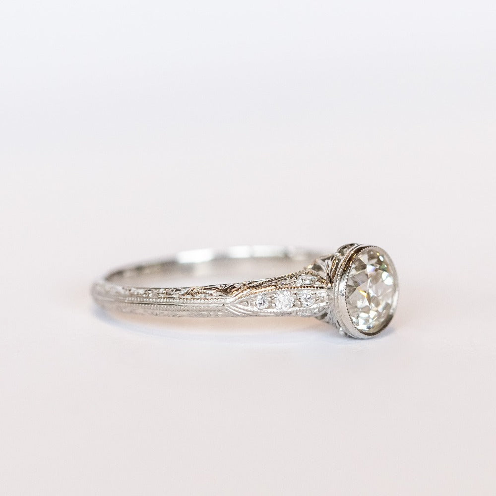 A platinum diamond engraved engagement ring with a bezel set old mine cut diamond center stone and a few diamond accents on each shoulder.