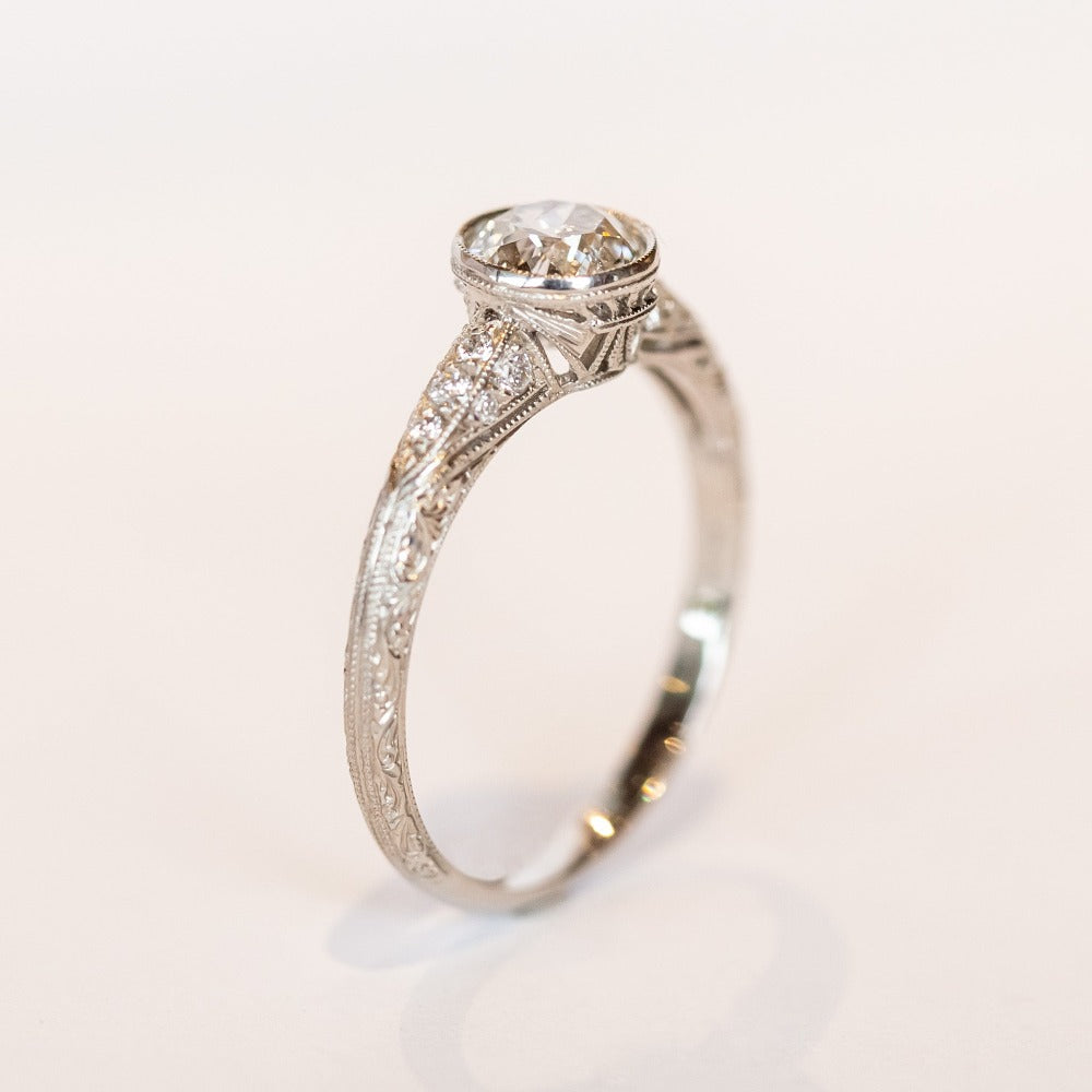 A platinum diamond engraved engagement ring with a bezel set old mine cut diamond center stone and a few diamond accents on each shoulder.