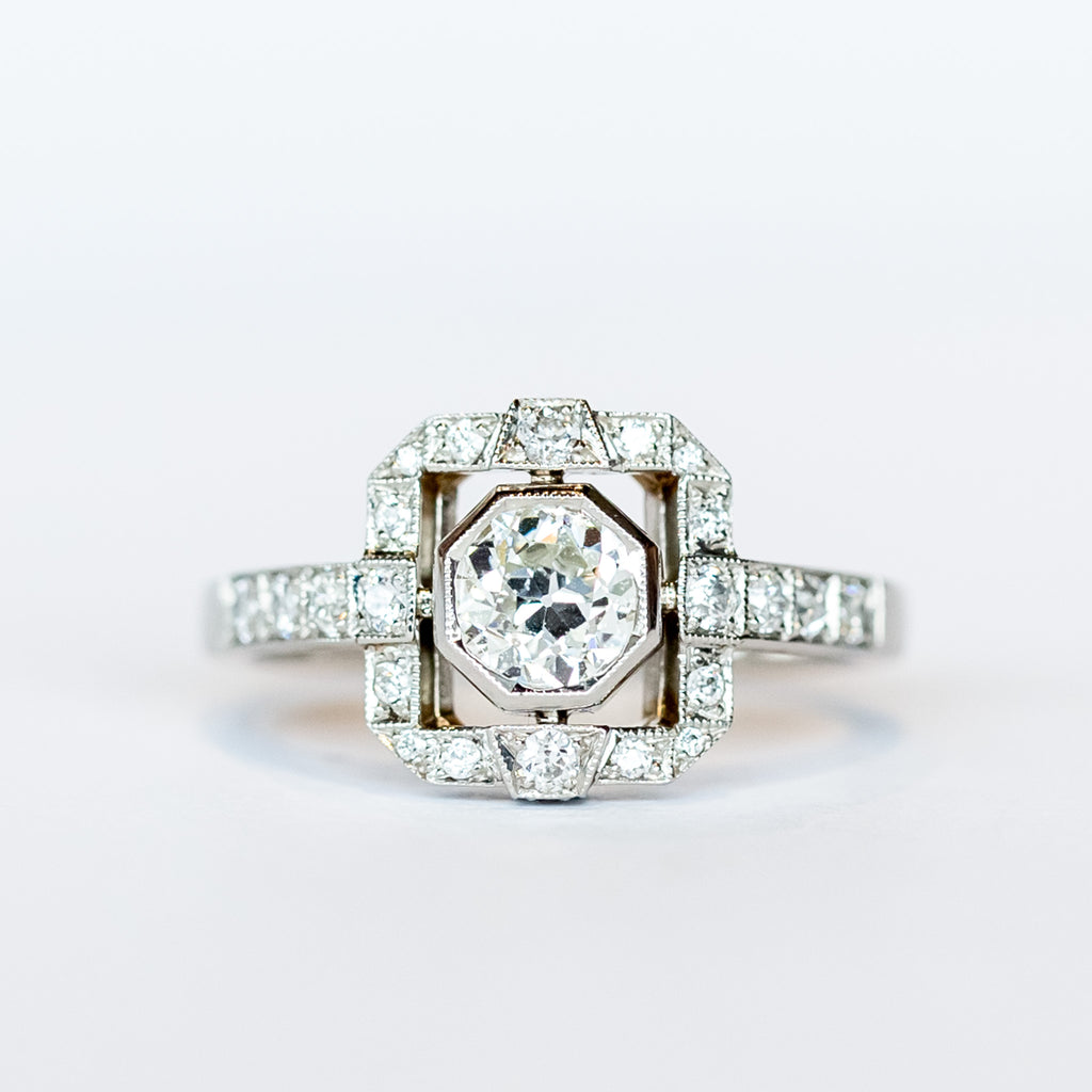 A platinum engagement ring featuring a bezel set central old European cut diamond framed by a square halo with milgrain details and diamond side stones with diamonds down each shoulder.