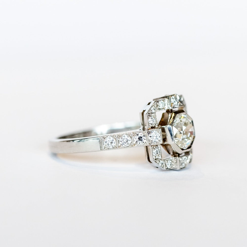 A platinum engagement ring featuring a bezel set central old European cut diamond framed by a square halo with milgrain details and diamond side stones with diamonds down each shoulder.