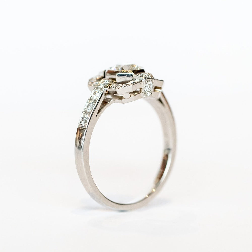 A platinum engagement ring featuring a bezel set central old European cut diamond framed by a square halo with milgrain details and diamond side stones with diamonds down each shoulder.