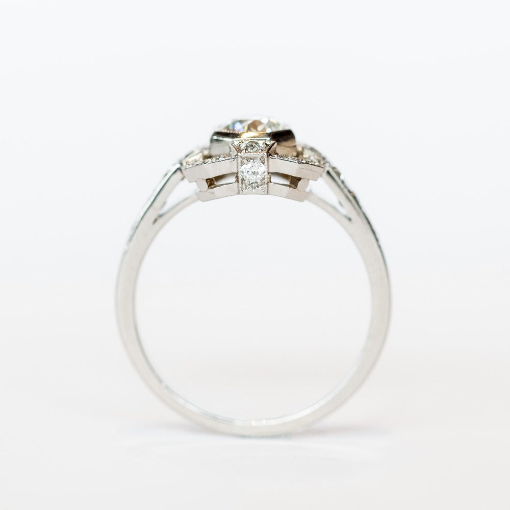 A platinum engagement ring featuring a bezel set central old European cut diamond framed by a square halo with milgrain details and diamond side stones with diamonds down each shoulder.