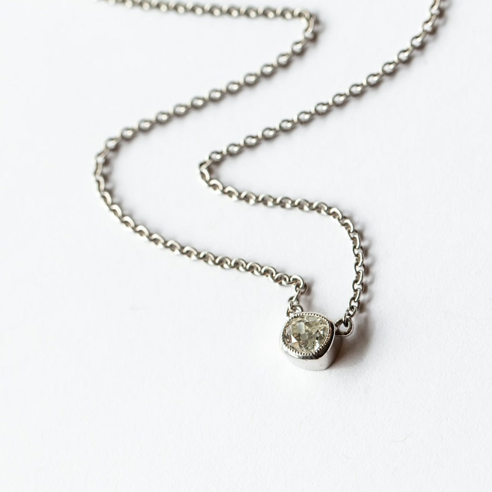 A cushion cut diamond in a platinum bezel setting with milgrain edge, stationed at the center of a platinum cable chain necklace.
