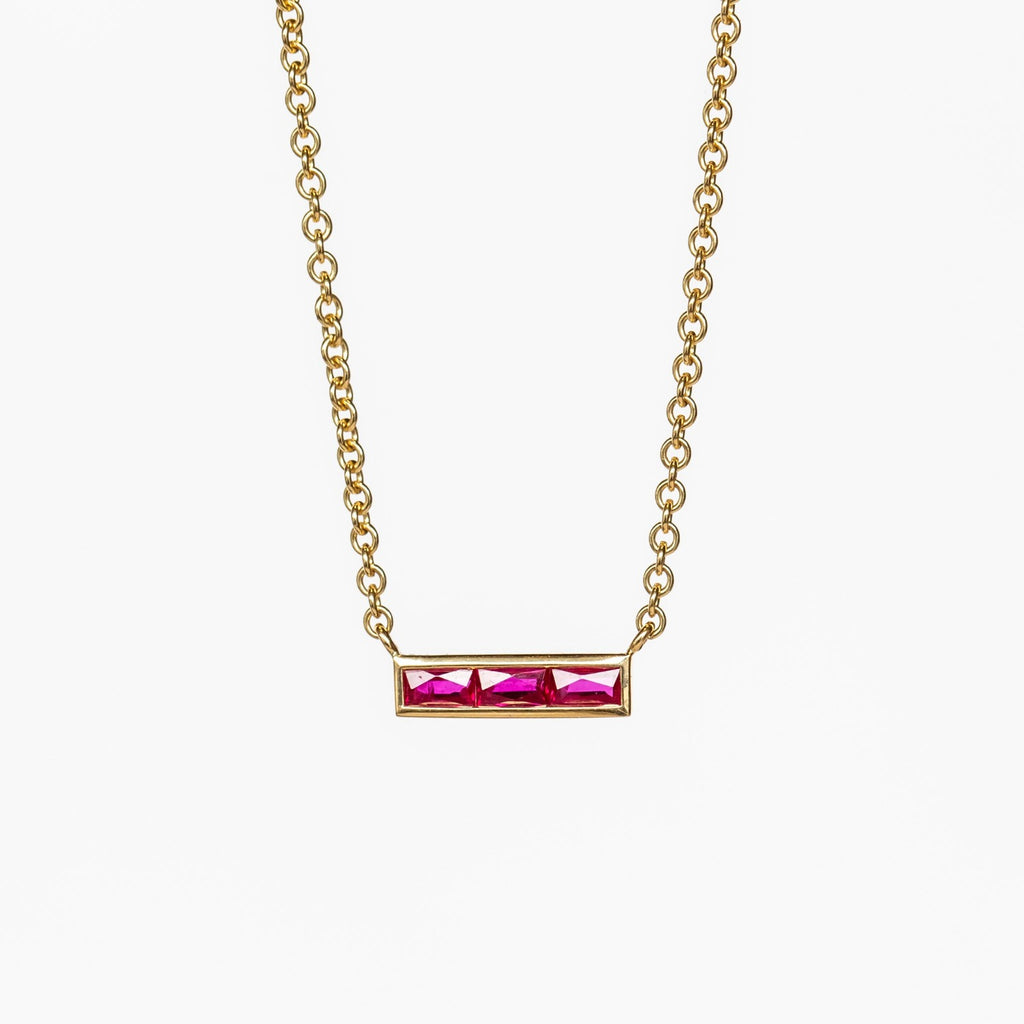 A horizontal bar gemstone necklace set with three French cut rubies, stationed at the center of a classic yellow gold cable chain necklace.