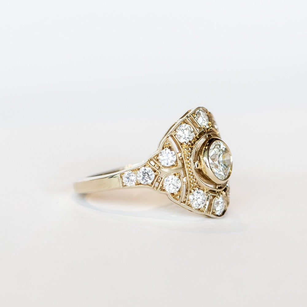 A vintage style diamond engagement ring with a bezel set center stone surrounded by an airy filigree setting and many other diamonds.