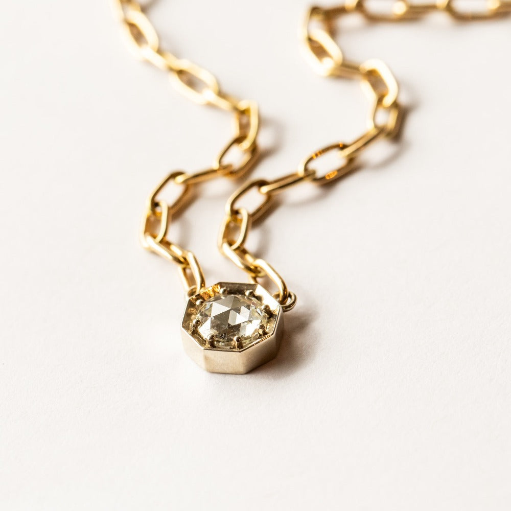 A rose cut diamond set in an octagonal white gold mounting centered on a yellow gold paperclip chain necklace.