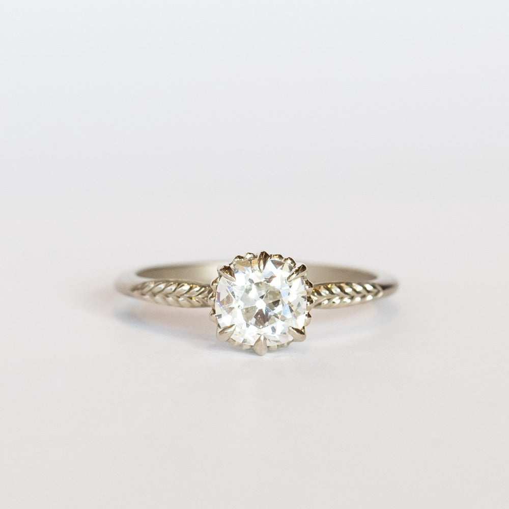 An old mine cut diamond is prong set into a 6-claw solitaire setting in white gold. The slender band has carved foliage motif on either side of the diamond.