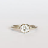 An old mine cut diamond is prong set into a 6-claw solitaire setting in white gold. The slender band has carved foliage motif on either side of the diamond.