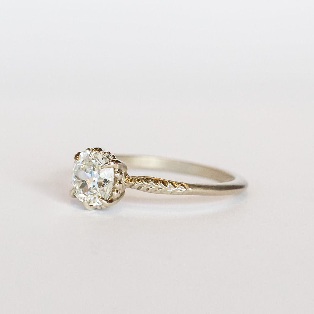 An old mine cut diamond is prong set into a 6-claw solitaire setting in white gold. The slender band has carved foliage motif on either side of the diamond.