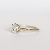 An old mine cut diamond is prong set into a 6-claw solitaire setting in white gold. The slender band has carved foliage motif on either side of the diamond.