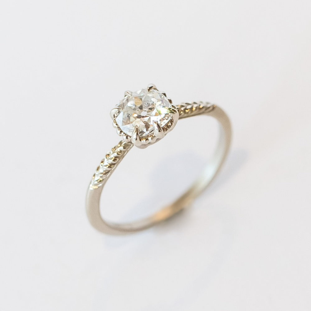 An old mine cut diamond is prong set into a 6-claw solitaire setting in white gold. The slender band has carved foliage motif on either side of the diamond.