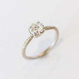 An old mine cut diamond is prong set into a 6-claw solitaire setting in white gold. The slender band has carved foliage motif on either side of the diamond.