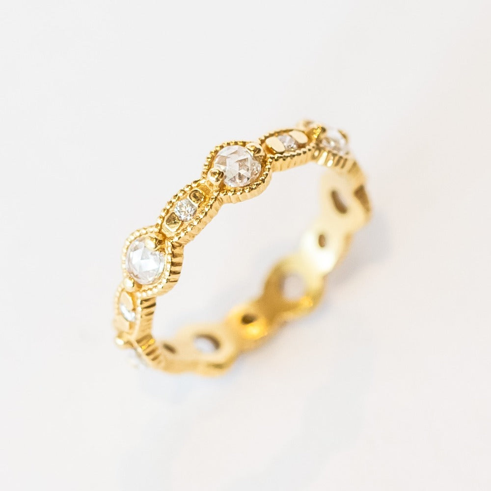 A yellow gold eternity band featuring round rose cut diamonds alternating with tiny brilliant round white diamonds. The alternating round and oval settings are accented with milgrain bed details.