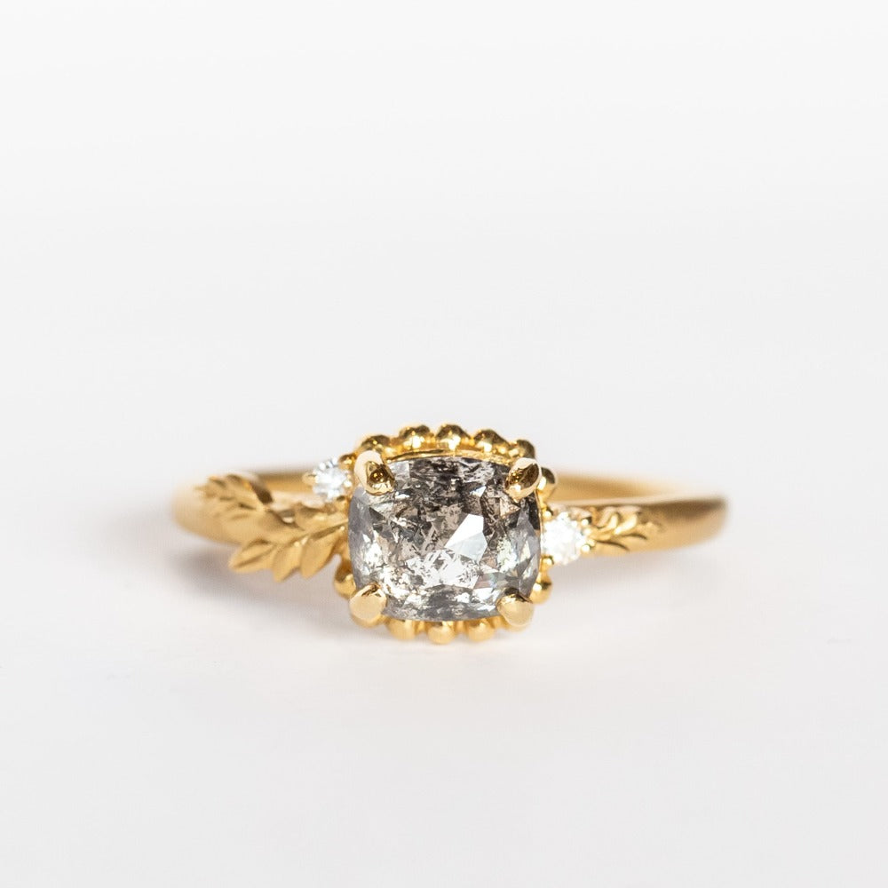 A cushion cut salt and pepper diamond is prong set into a yellow gold engagement ring. The sides of the setting feature an asymmetrical floral motif set with two tiny accent diamonds.