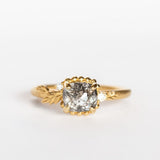 A cushion cut salt and pepper diamond is prong set into a yellow gold engagement ring. The sides of the setting feature an asymmetrical floral motif set with two tiny accent diamonds.