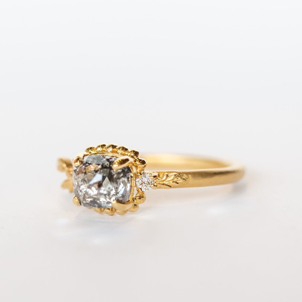 A cushion cut salt and pepper diamond is prong set into a yellow gold engagement ring. The sides of the setting feature an asymmetrical floral motif set with two tiny accent diamonds.