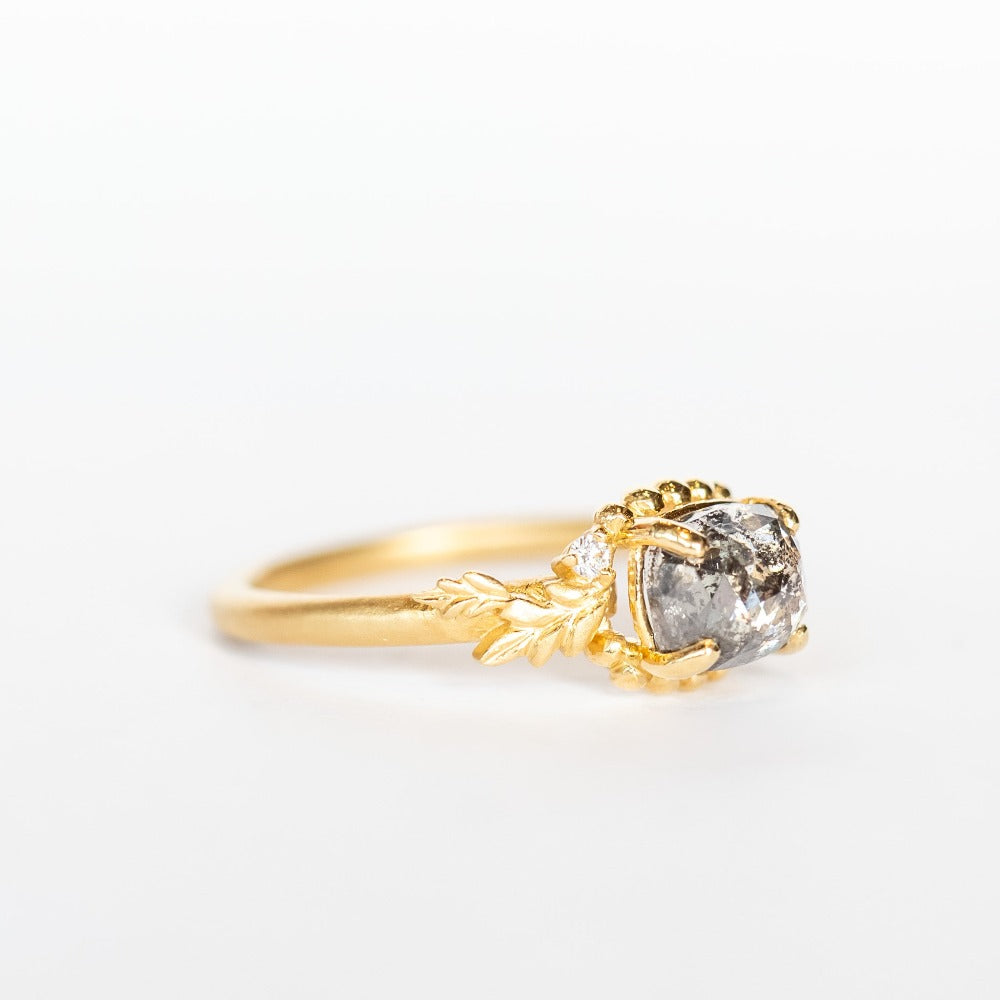 A cushion cut salt and pepper diamond is prong set into a yellow gold engagement ring. The sides of the setting feature an asymmetrical floral motif set with two tiny accent diamonds.