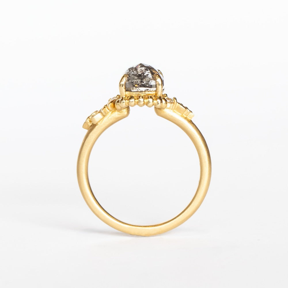 A cushion cut salt and pepper diamond is prong set into a yellow gold engagement ring. The sides of the setting feature an asymmetrical floral motif set with two tiny accent diamonds.