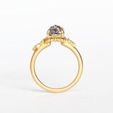 A cushion cut salt and pepper diamond is prong set into a yellow gold engagement ring. The sides of the setting feature an asymmetrical floral motif set with two tiny accent diamonds.