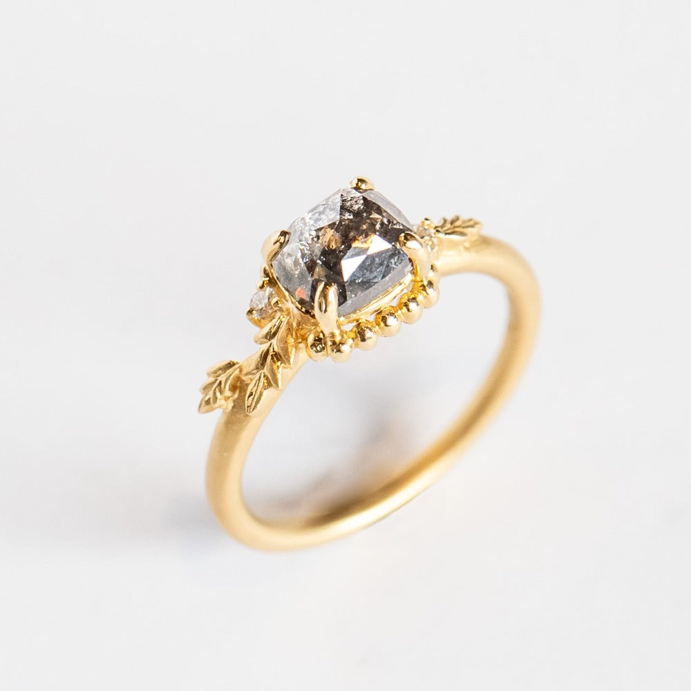 A cushion cut salt and pepper diamond is prong set into a yellow gold engagement ring. The sides of the setting feature an asymmetrical floral motif set with two tiny accent diamonds.