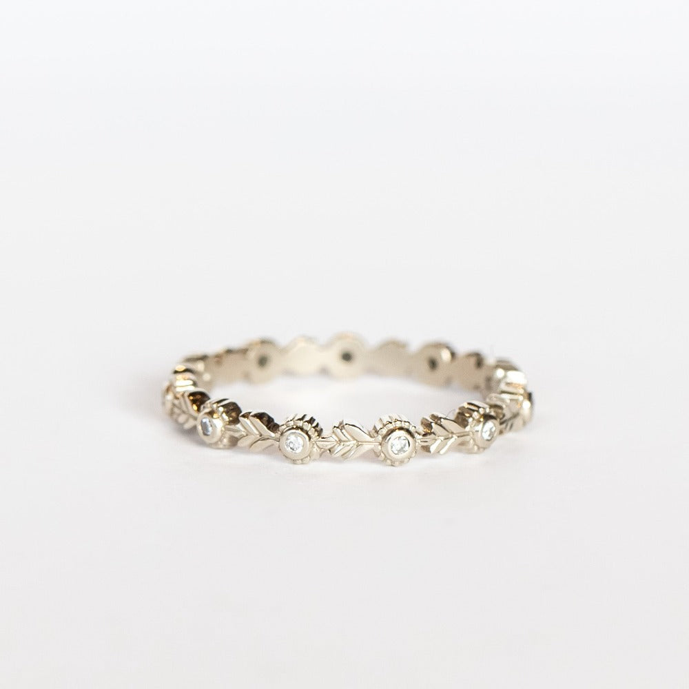 A white gold eternity wedding band featuring leaf patterns alternating with tiny bezel set diamonds surrounded with milgrain dot accents.
