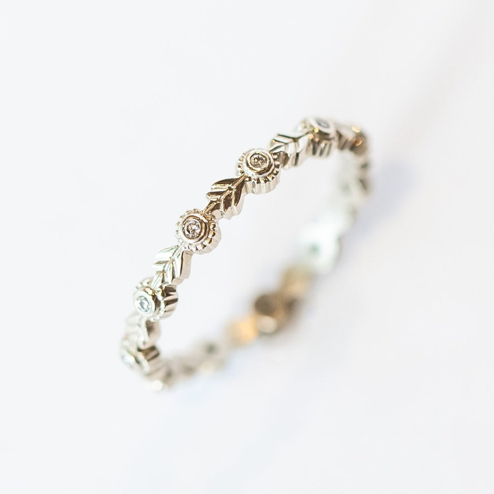 A white gold eternity wedding band featuring leaf patterns alternating with tiny bezel set diamonds surrounded with milgrain dot accents.