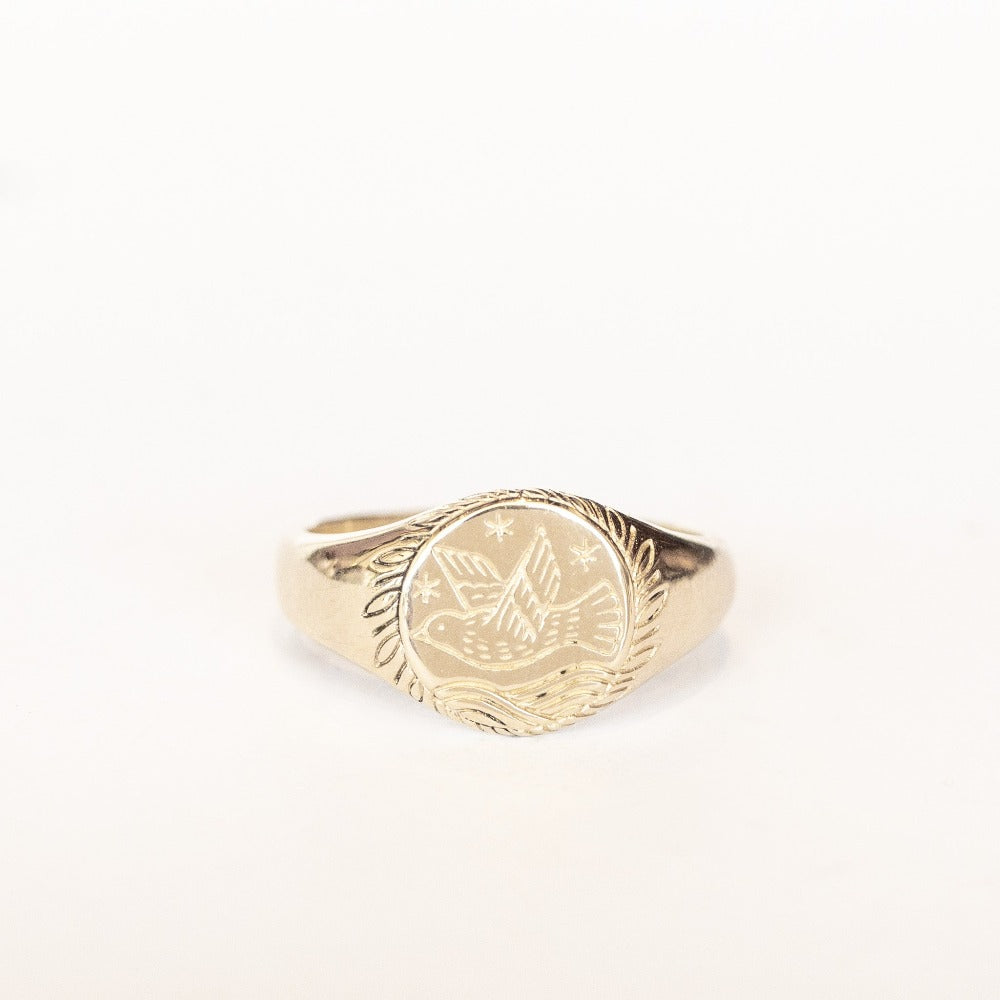 A gold signet style pinkie ring featuring an engraved bird, starry sky, and ocean surrounded by leaves.