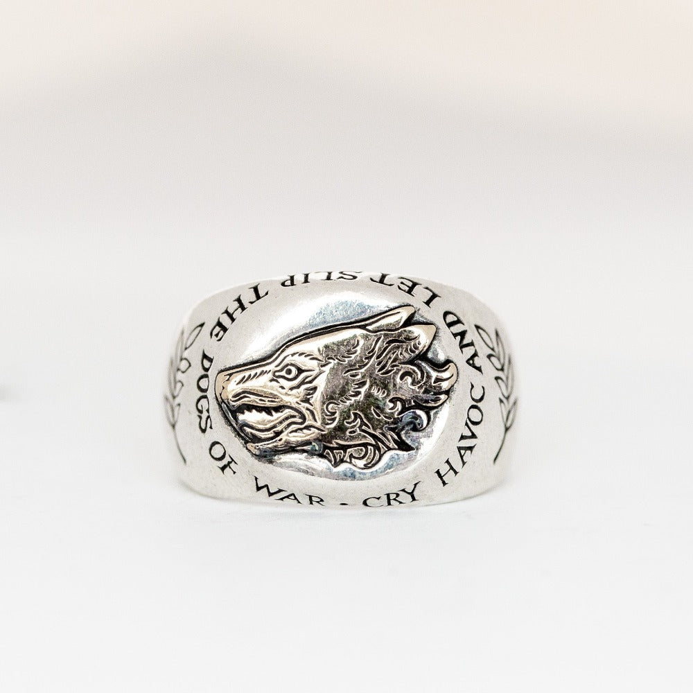 A chunky silver signet ring with gold inlaid dog's head. It is engraved with the words: 'Cry havoc and let slip the dogs of war', a quote from William Shakespeare's Julius Caesar. 