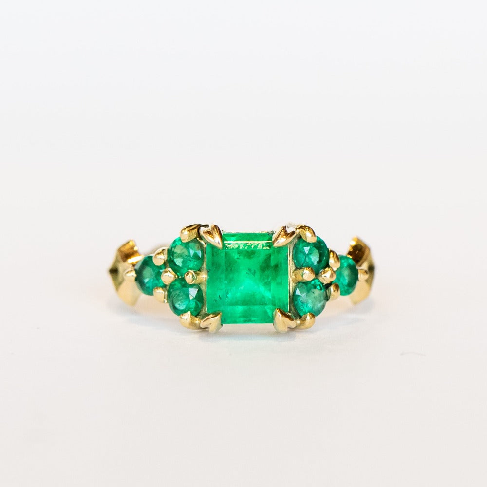 An emerald ring set of engagement ring and wedding band. Features an emerald cut emerald gemstone center flanked by six round emeralds, and a graduating size emerald band that contours to fit.
