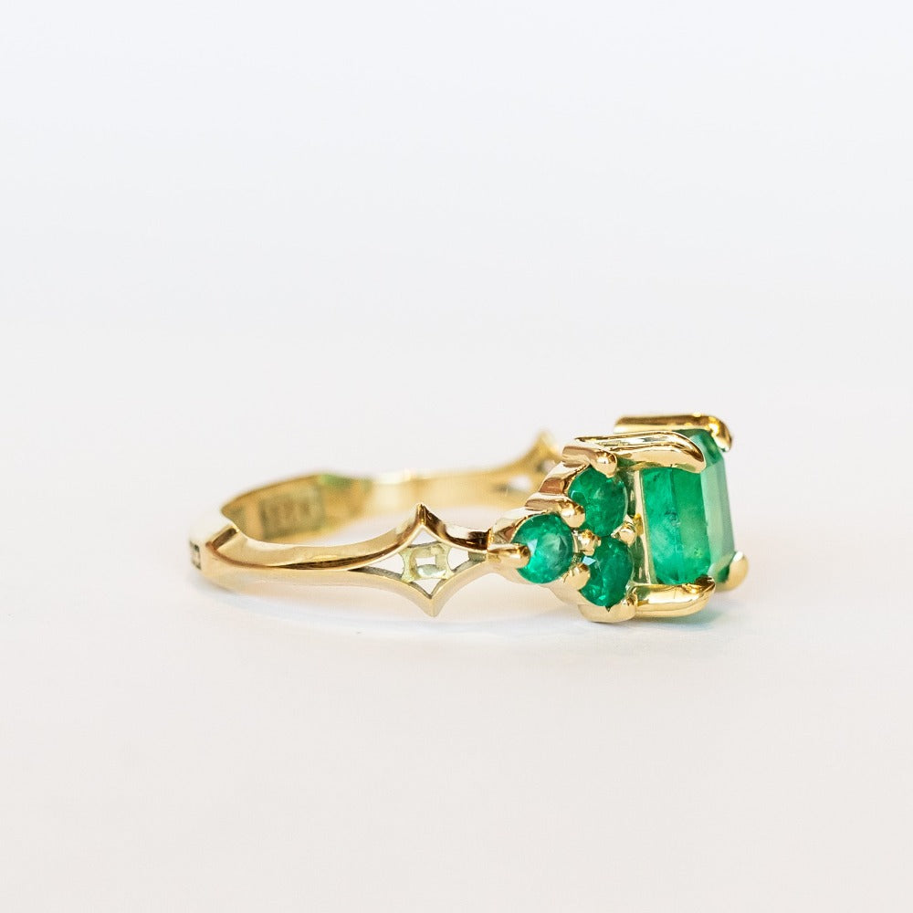 An emerald ring set of engagement ring and wedding band. Features an emerald cut emerald gemstone center flanked by six round emeralds, and a graduating size emerald band that contours to fit.